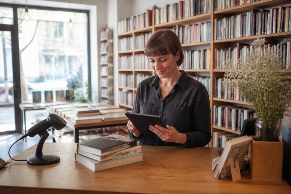 Una libraia indipendente al bancone che gestisce le vendite con un tablet, scaffali di libri sullo sfondo, un lettore di codici a barre vicino alla cassa, luce naturale dalla vetrina