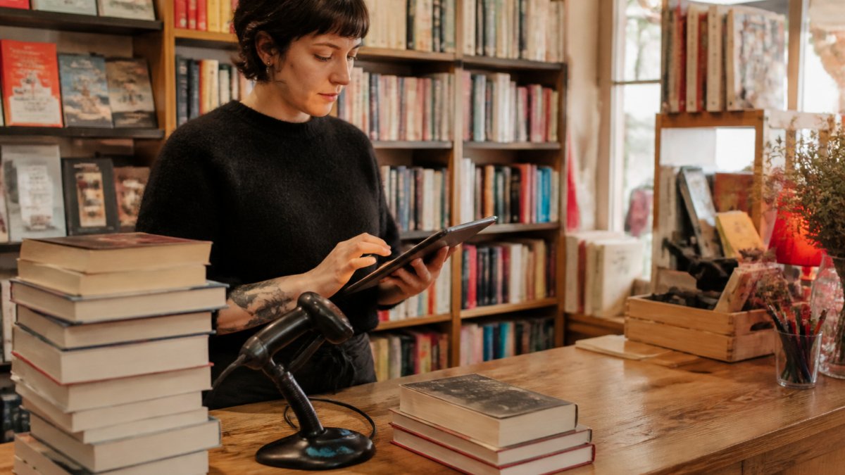 A bookseller at a wooden counter using a tablet as a point of sale, with stacks of books and a barcode scanner nearby in a warm independent bookstore