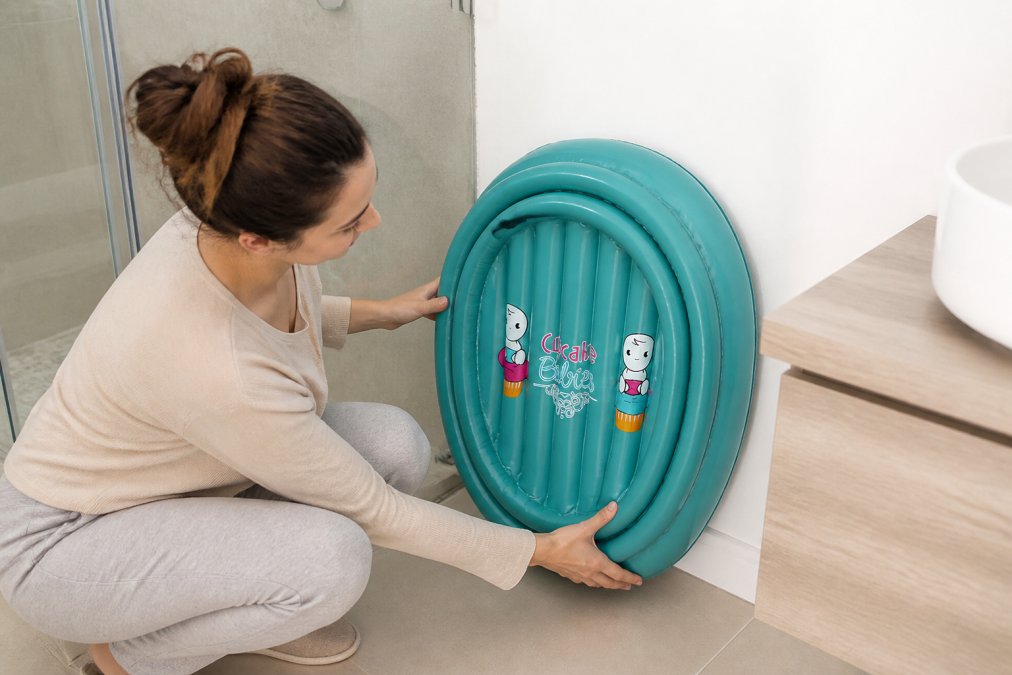Parent pliant une baignoire gonflable compacte dans un petit espace de salle de bain, montrant le rangement vertical contre un mur blanc et la facilité de manipulation
