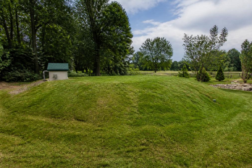 A grassy mound septic system in a rural backyard with a small pump housing, surrounded by trees and open sky, showing the elevated drainfield above the natural soil