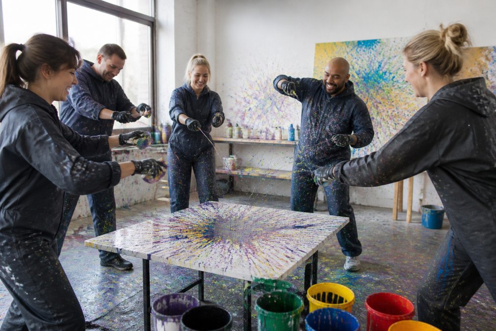 Eine diverse Gruppe von Mitarbeitern beim Action Painting Workshop, die gemeinsam bunte Kunstwerke mit Schleuderbildern erschaffen, mit fröhlichen Gesichtern und kreativer Energie in einem hellen Studio