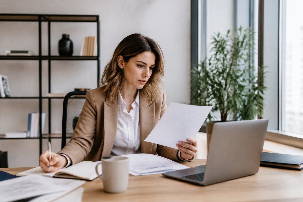 Junge Unternehmerin sitzt am Schreibtisch mit Laptop, Notizbuch und Tasse Kaffee, studiert Dokumente zur Unternehmensgründung in hellem, modernem Büro