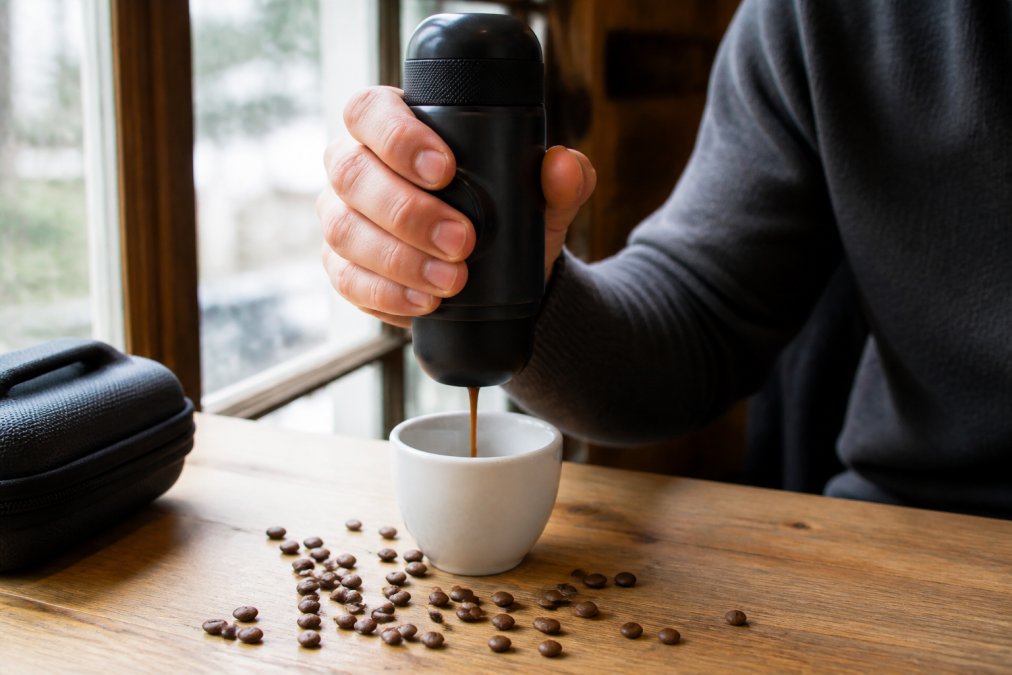 En person holder en håndholdt Minipresso espressomaskine mens den brygger kaffe over en hvid kop på et træbord med kaffebønner spredt omkring, naturligt dagslys fra vinduet