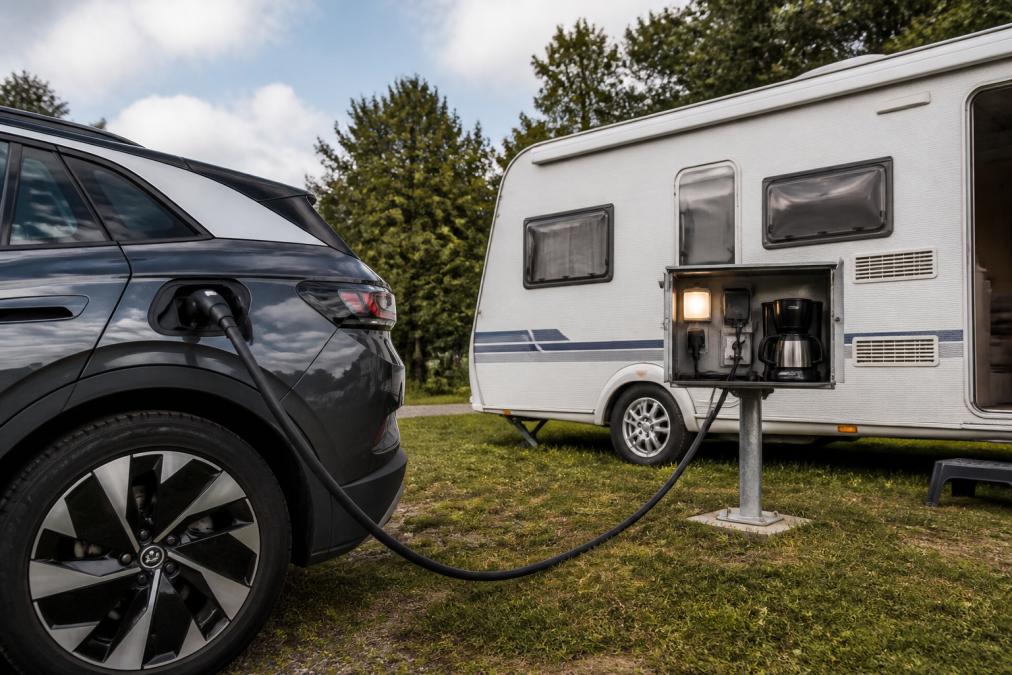 En elbil parkeret ved en campingvogn på en dansk campingplads. Et kabel løber fra bilens Type 2-stik til et eksternt strømskab ved campingvognen, hvor en lille lampe og en kaffemaskine er tilsluttet.
