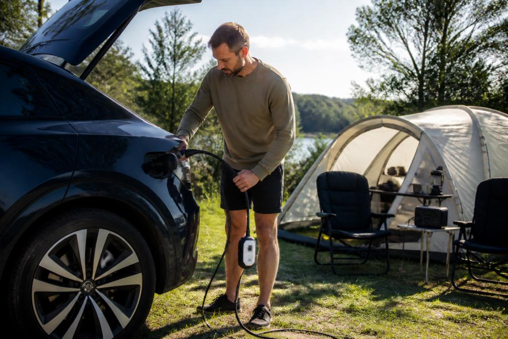 A person next to an electric car at a campsite connecting V2L equipment to an external power consumer, demonstrating practical application in daylight with a camping tent in the background