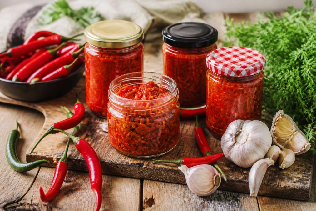 Arrangement artistique de pots de pâtes de piments avec étiquettes visibles, entourés de piments frais, ail et herbes aromatiques sur une table en bois rustique avec éclairage naturel