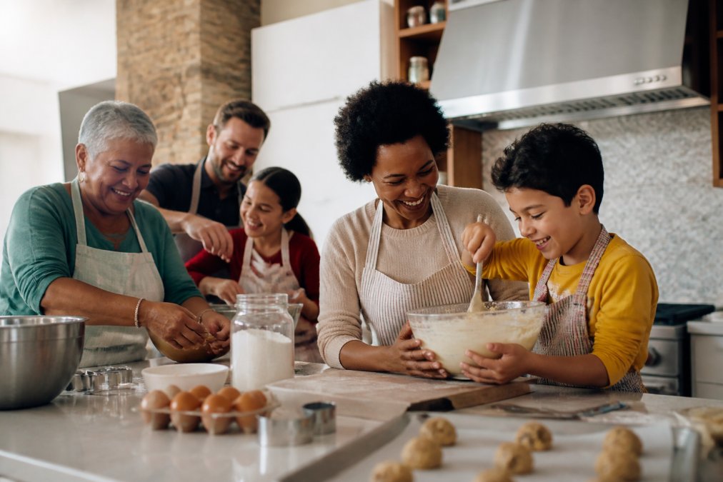 A diverse group of home bakers in a bright home kitchen, including Black, white, and Latino people of different ages, mixing cookie dough and laughing. One woman holds a bowl of Lura's Kitchen cookie mix while a child stirs beside her. Warm natural light, candid and lifelike.