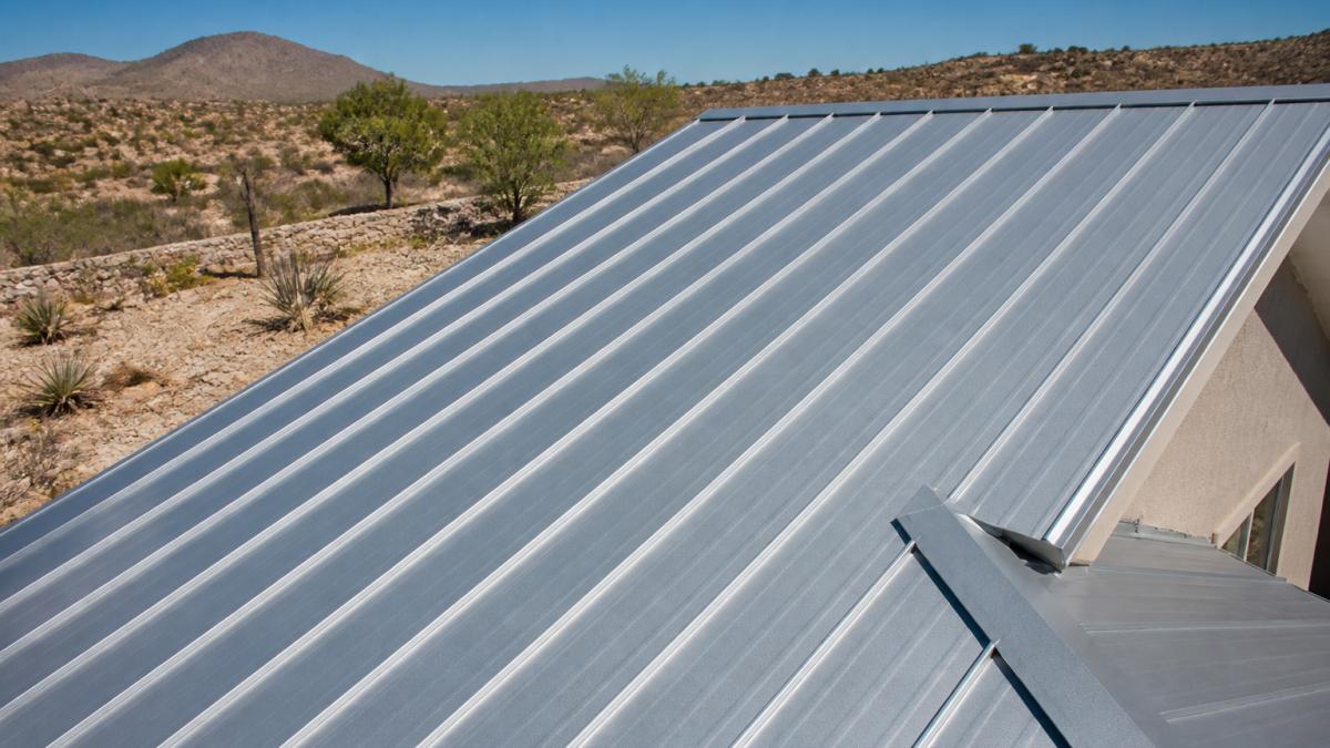 Close-up of standing seam metal roof installed on residential home in desert climate, showing reflective silver surface and seam details with clear blue sky and desert landscape in background