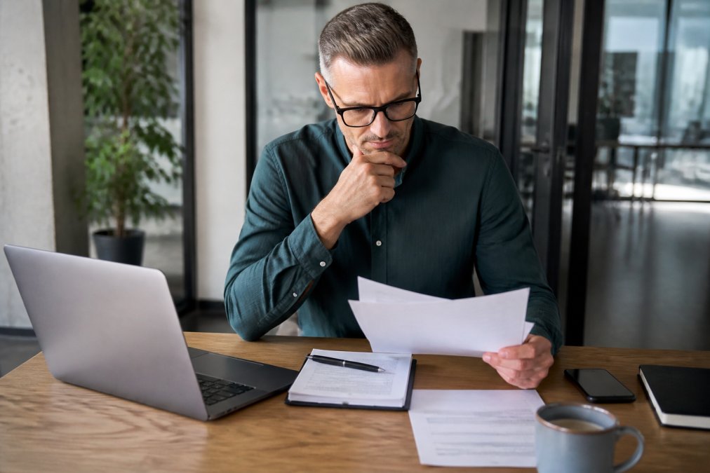 A business professional comparing documents on a laptop and notepad in a bright office, with a coffee cup nearby, looking thoughtful