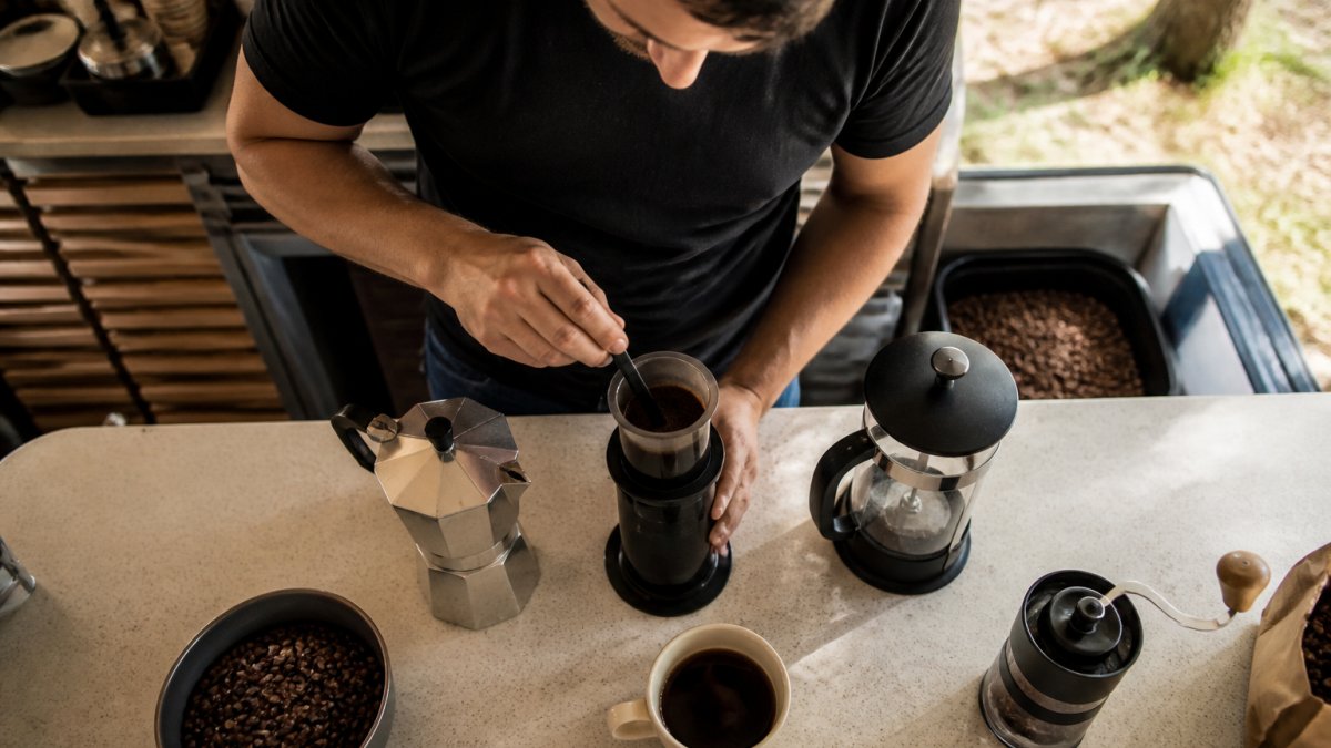 Overhead view of a moka pot, AeroPress, and French press arranged on a kitchen counter with whole coffee beans, a burr grinder, and a cup of brewed coffee