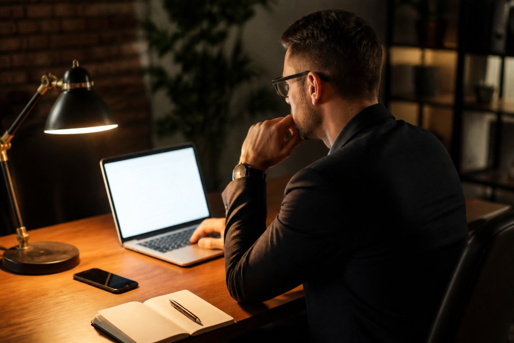 Sales manager reviewing automated follow-up sequence on a laptop screen in a dim office, phone and notebook on desk, warm desk lamp lighting