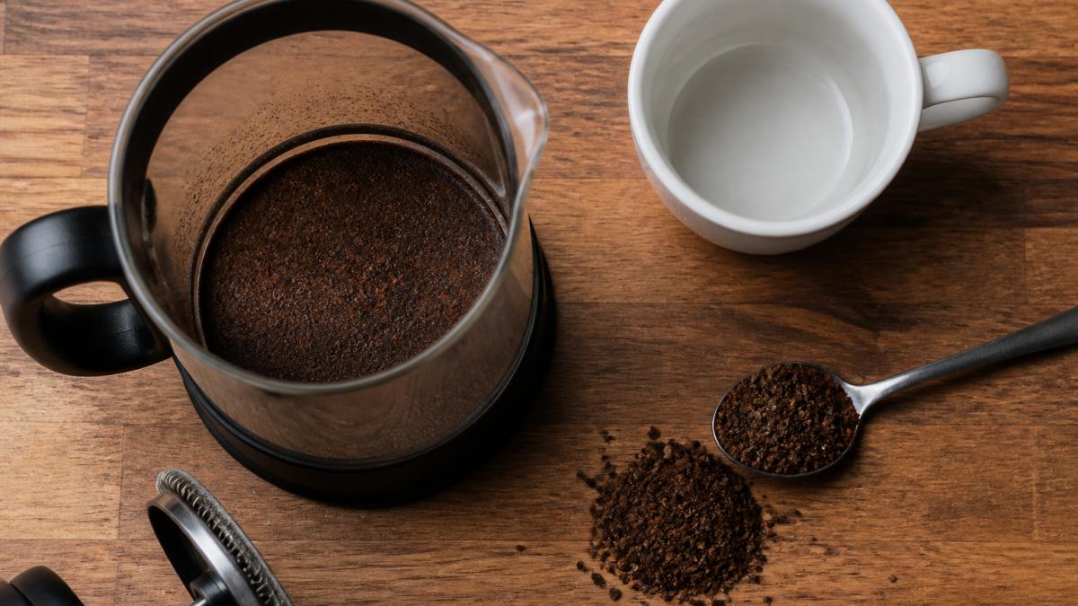 French press on a wooden counter with fine grounds in the carafe next to a ceramic mug and a spoon of coffee
