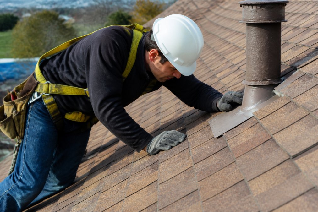 Professional roofer in safety harness and hard hat inspecting roof surface near metal flashing, examining seams and penetration points with close attention