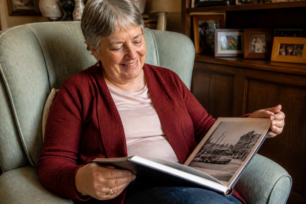 An older adult holding a nostalgic photo book, smiling while looking at a page with black-and-white historical photos, seated in a comfy armchair in a warm living room