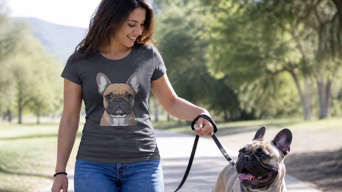 A woman in her thirties wearing a fitted graphic t-shirt with a French Bulldog illustration, standing outdoors with her dog on a leash, natural daylight, casual setting