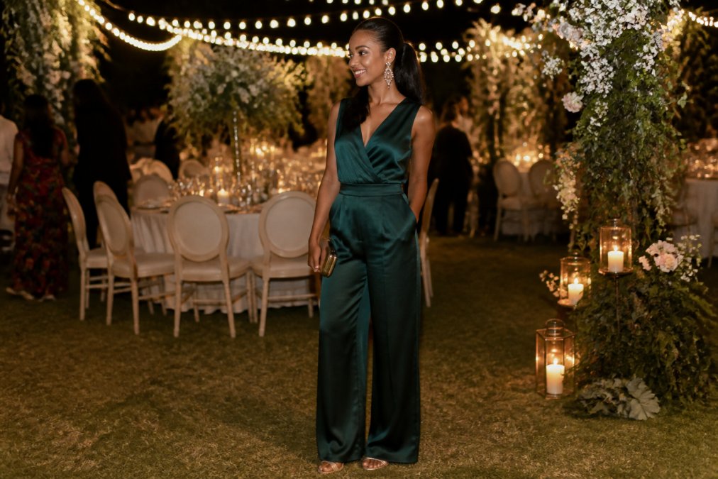 A stylishly dressed woman in a tailored wide-leg silk jumpsuit in deep jewel tones, wearing statement chandelier earrings and strappy heeled sandals, standing at an outdoor wedding reception with floral arrangements and string lights in the background