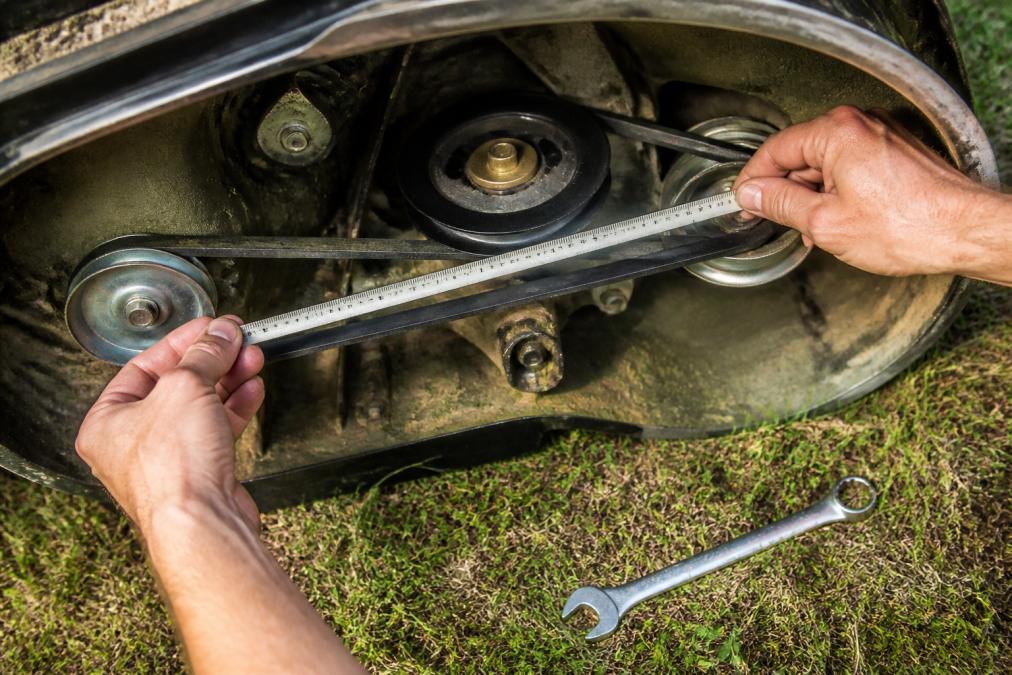 Close-up of hands using a flexible tape measure around a lawn mower deck belt still routed on the pulleys, with a wrench on the grass nearby