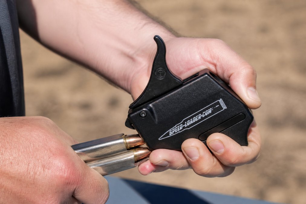Close-up of a shooter's hands using a metal speed loader at an outdoor range, showing the mechanical advantage and ergonomic grip in action with natural sunlight