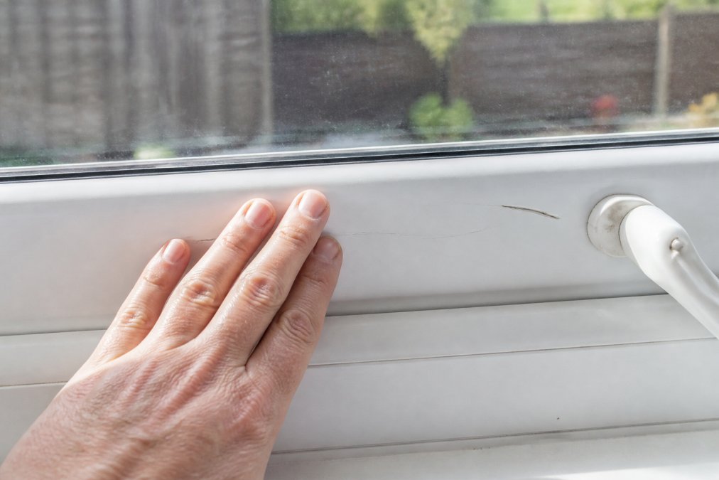 Close-up of a homeowner's hand running along a white uPVC window frame, with visible fine scratches catching the daylight beside a deeper gouge near the handle