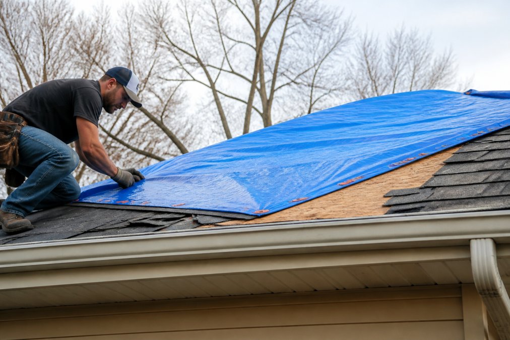 Professional roofer securing heavy-duty waterproof tarp over storm-damaged roof section with visible missing shingles and exposed decking, using proper fastening techniques in daylight
