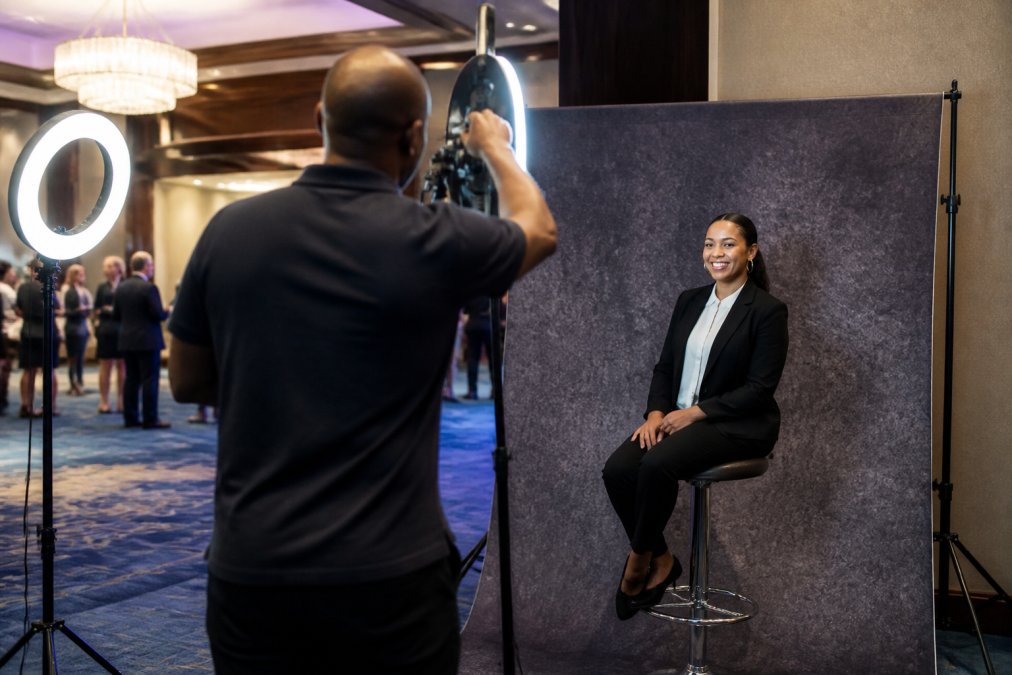Professional photographer adjusting lighting setup at a branded photo booth station with custom backdrop, LED ring lights, and a guest smiling at the camera