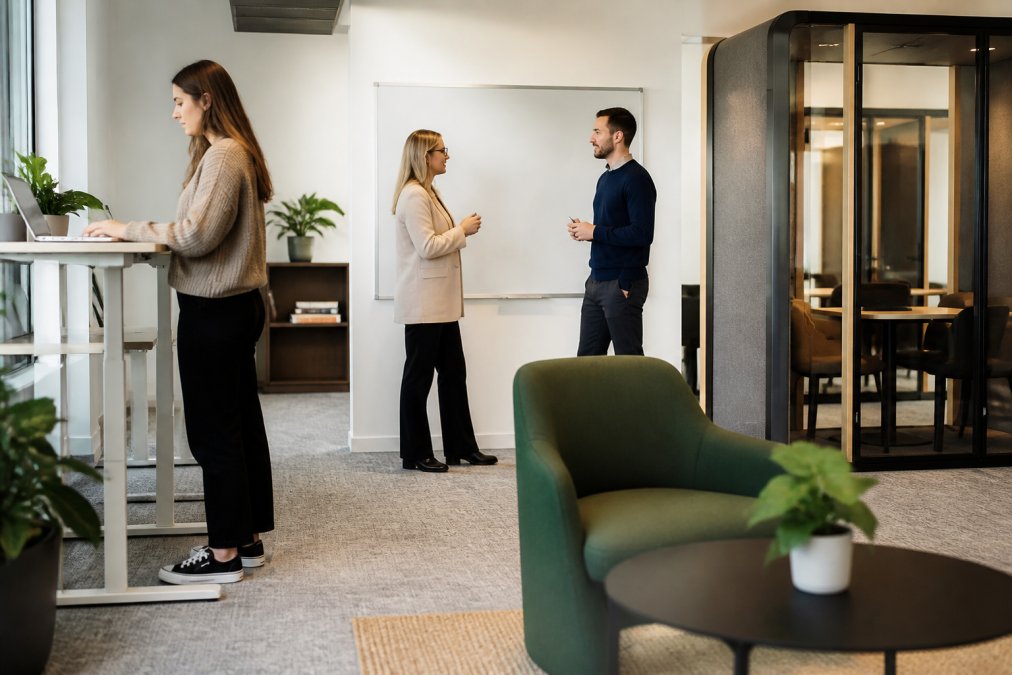 A bright modern office with a mix of standing desks, soft seating and a small meeting pod, two people talking near a whiteboard and one person working at a standing desk