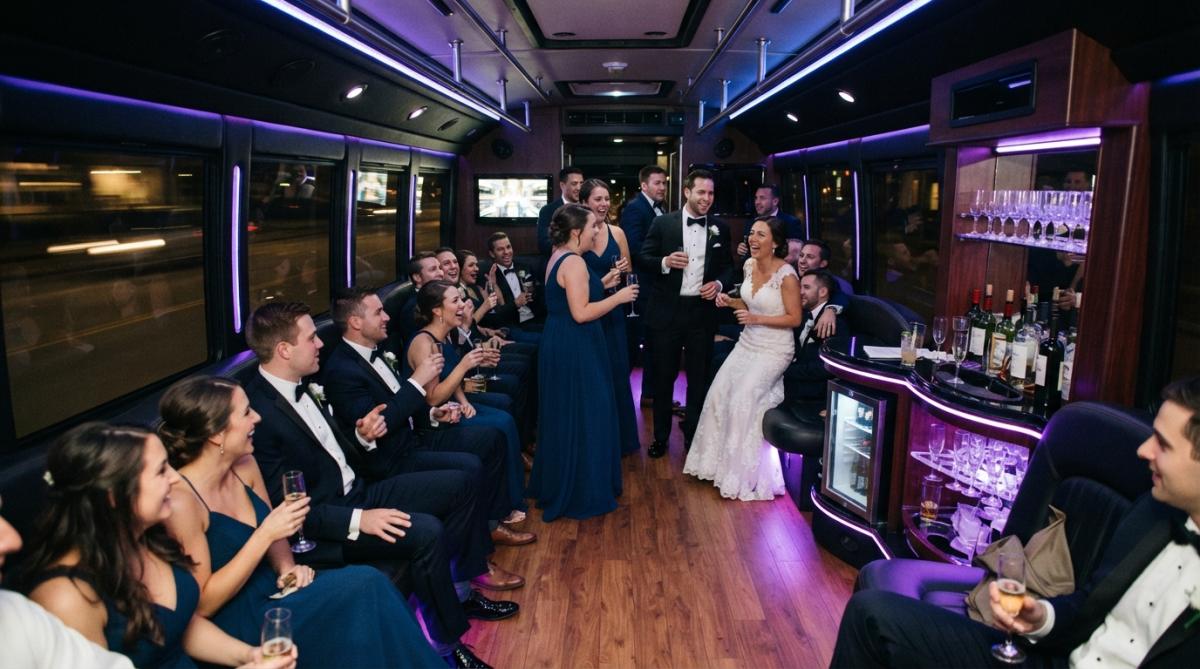 Luxury party bus interior with guests in formal wedding attire socializing, ambient purple and blue LED lighting, comfortable perimeter seating, spacious dance floor area in center