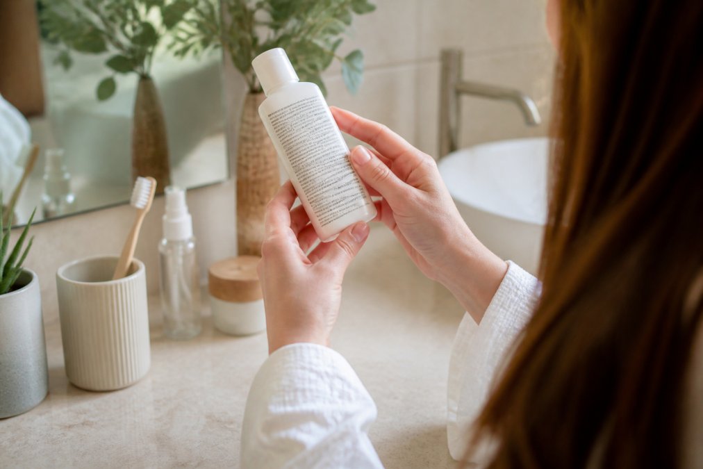 Woman holding a skincare bottle and reading the ingredient label on a bright bathroom counter