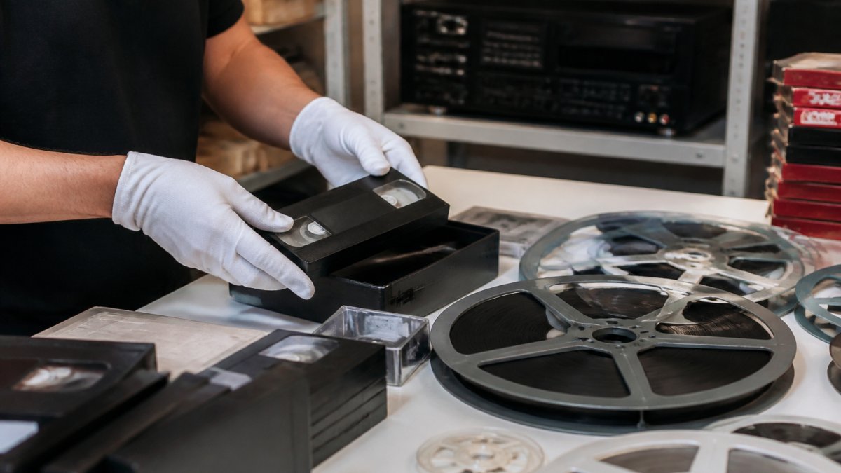 Professional carefully handling vintage VHS tapes and film reels on a work table with professional digitisation equipment visible in the background, hands wearing cotton gloves