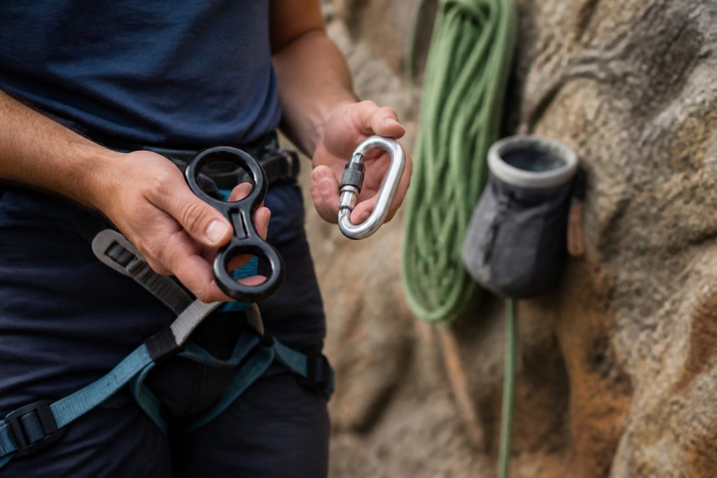 Climber holding a figure-eight descender and locking carabiner alongside various belay devices on a rock face