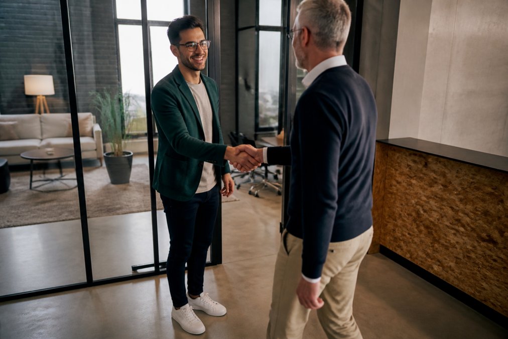 A confident startup founder in a relaxed blazer and clean sneakers shaking hands with an investor in a modern office with glass walls and natural light