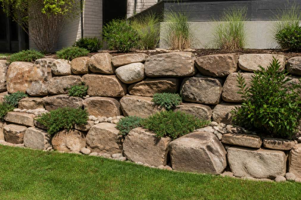 Completed natural stone retaining wall in residential backyard with stacked boulders creating rustic landscape feature, showing finished aesthetic and natural drainage between stones