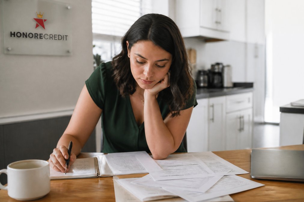 A person at a kitchen table reviewing credit reports and financial documents on a laptop, with a notebook and coffee nearby, looking focused and determined