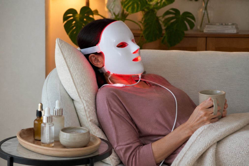 A woman in her forties relaxing on a sofa wearing a hands-free LED facial mask, with a small tray of serum bottles and a clay mask jar on the side table beside her