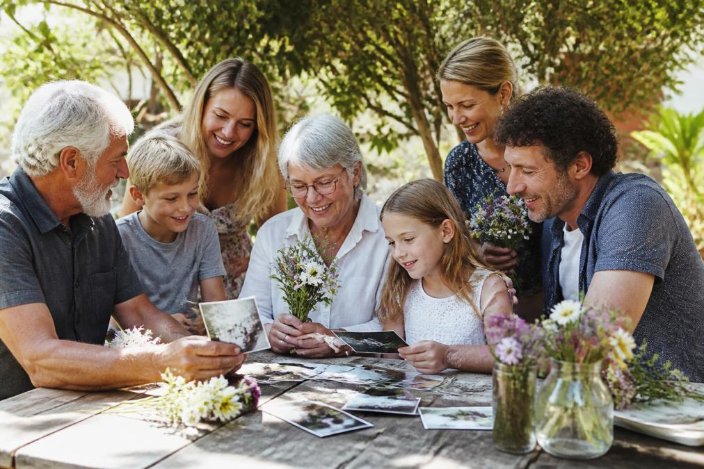 Family members of different ages gathered outdoors in a sunlit garden setting, sharing stories and smiling together while holding flowers and looking through old photographs on a picnic table