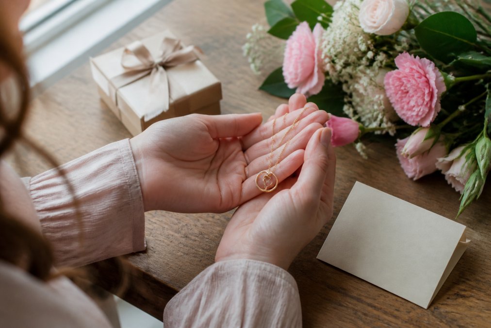 A woman's hands gently holding a delicate gold necklace with a small personalized message card resting beside it on a wooden table, soft natural window light