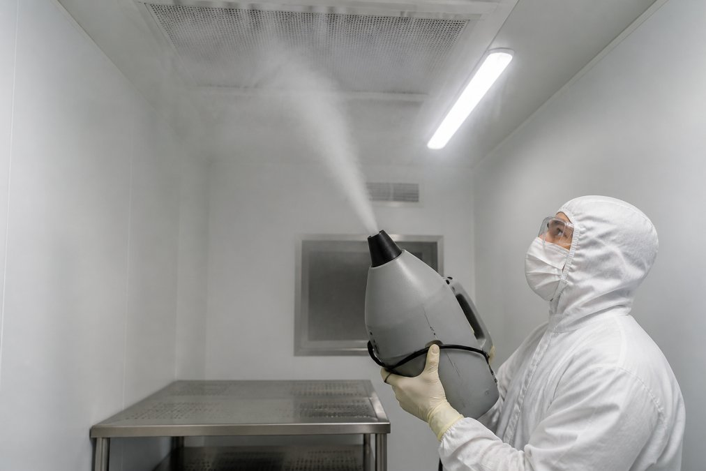 Process engineer releasing visible aerosol tracer into cleanroom environment to observe airflow patterns around work surfaces and HEPA filter returns under bright cleanroom lighting