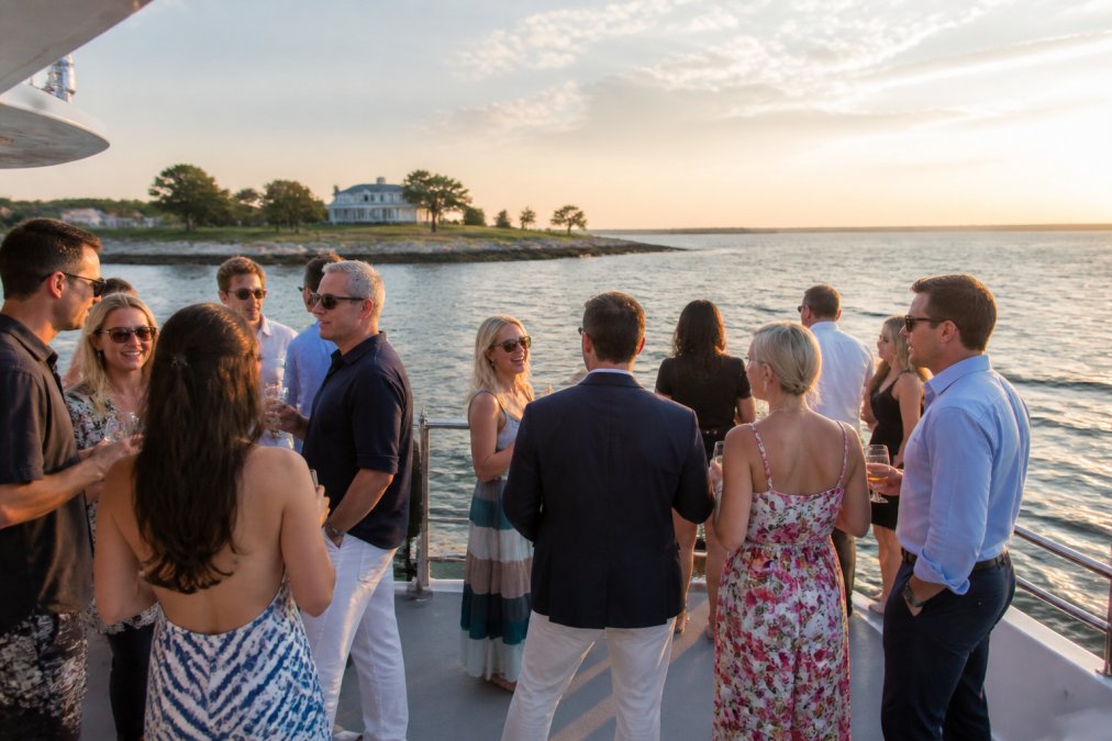 A private charter boat on Narragansett Bay with guests gathered on the open deck, water and rocky shoreline visible in the background, sunset lighting casting golden tones, casual celebration atmosphere with people mingling