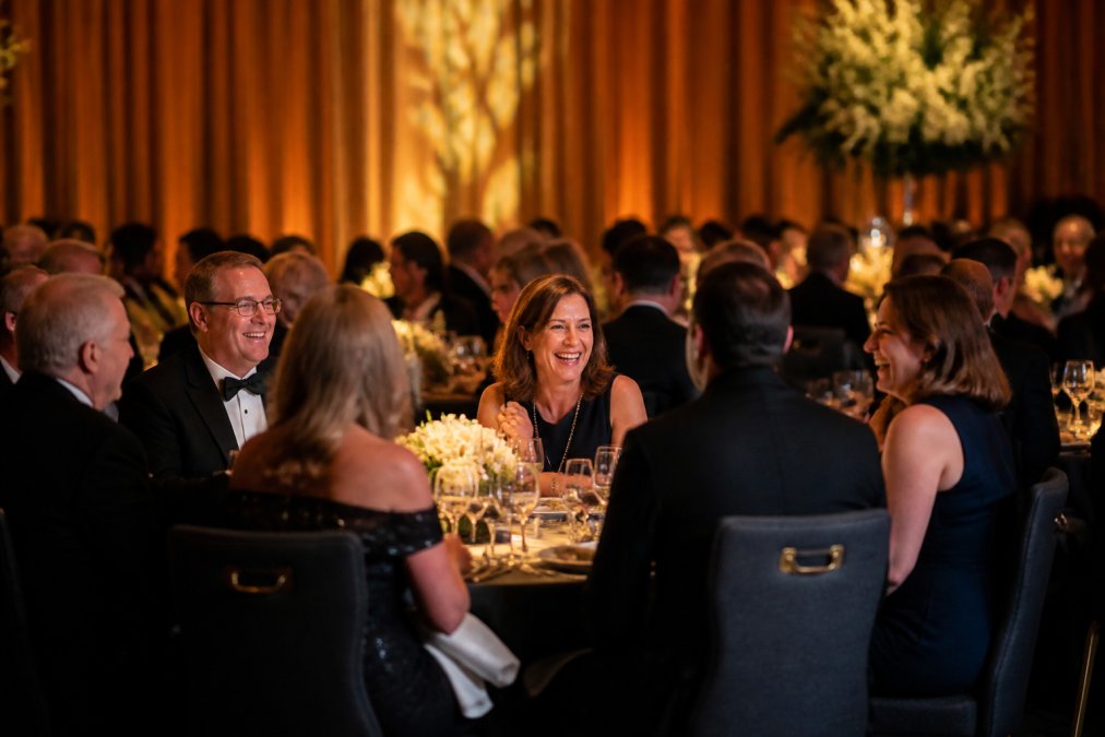 Professional event photographer discreetly capturing candid moments of attendees laughing and conversing at a corporate gala, using a prime lens from an unobtrusive position, warm stage lighting illuminating the scene