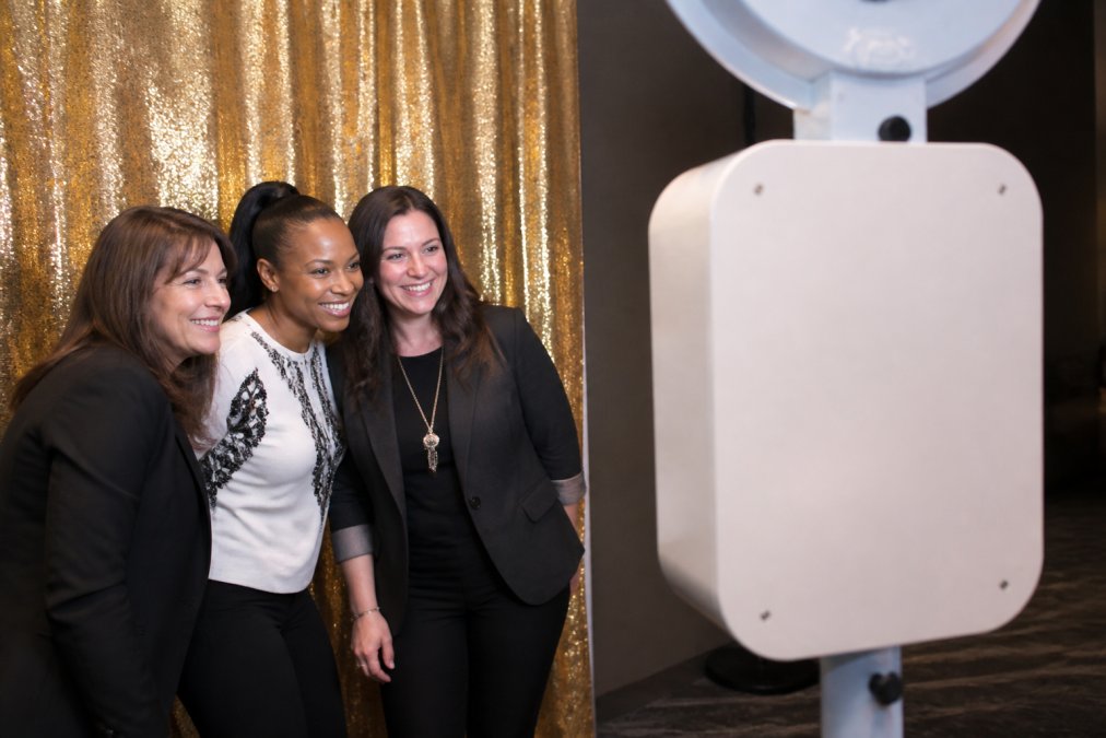 Guests smiling and posing in front of an AI photo booth displaying colorful digital overlays and branded backdrop at a corporate event, with instant digital transformation visible on the booth's display screen