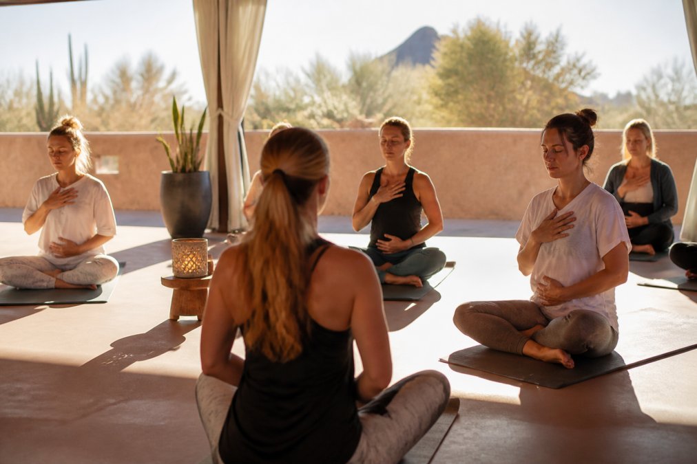 A small group of yoga practitioners seated on mats in a sunlit desert studio, practicing guided breathwork during an immersive retreat session