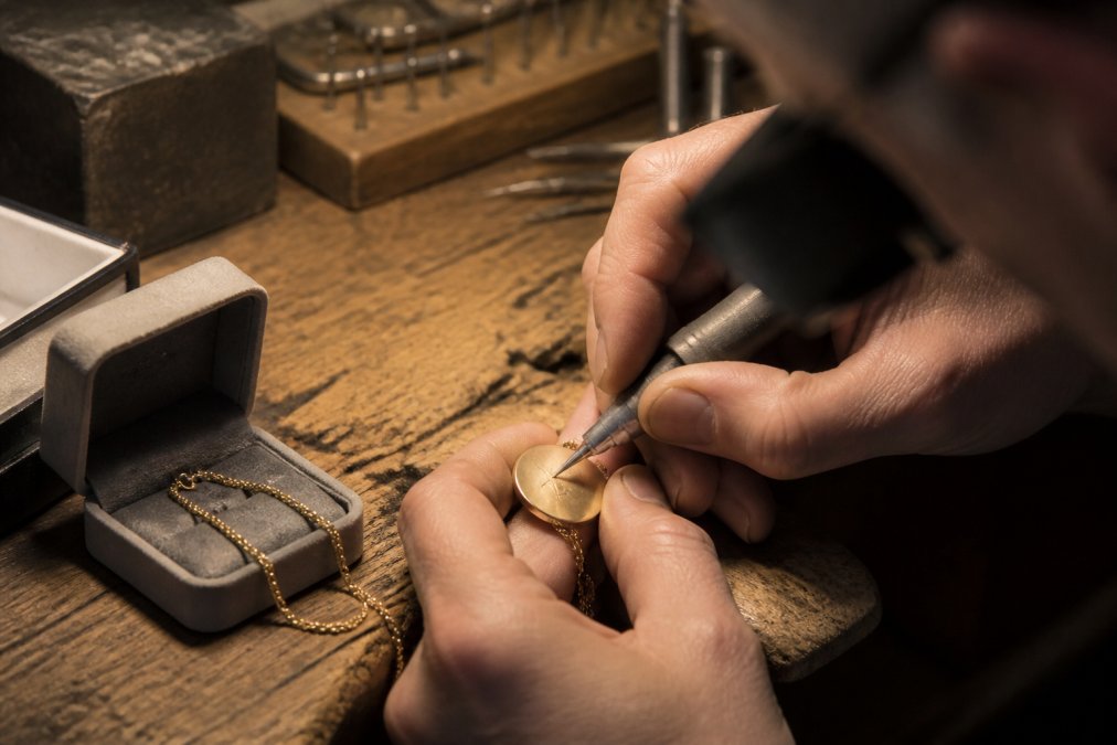 Close-up of an artisan's hands engraving a message onto a gold necklace pendant at a wooden workbench, small tools and a soft gift box nearby, warm workshop lighting