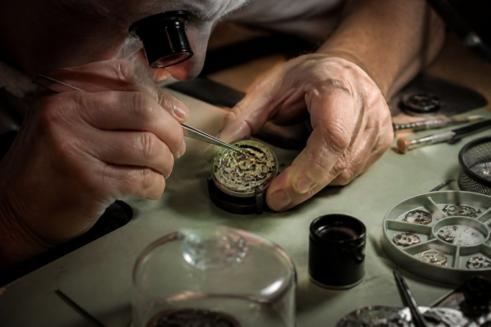 Close-up of a watchmaker's hands using tweezers to assemble an automatic watch movement at a clean workbench, with small tools and watch components visible under warm task lighting