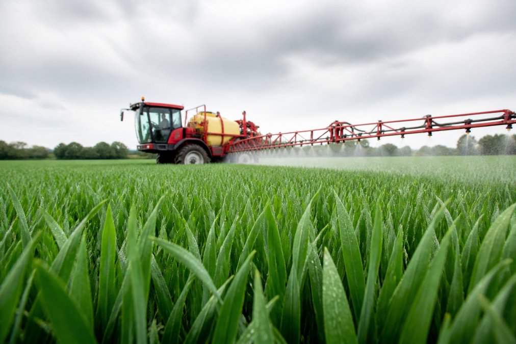 A crop sprayer applying foliar treatment to a field of winter wheat under an overcast sky, with droplets visible on the leaves in the foreground
