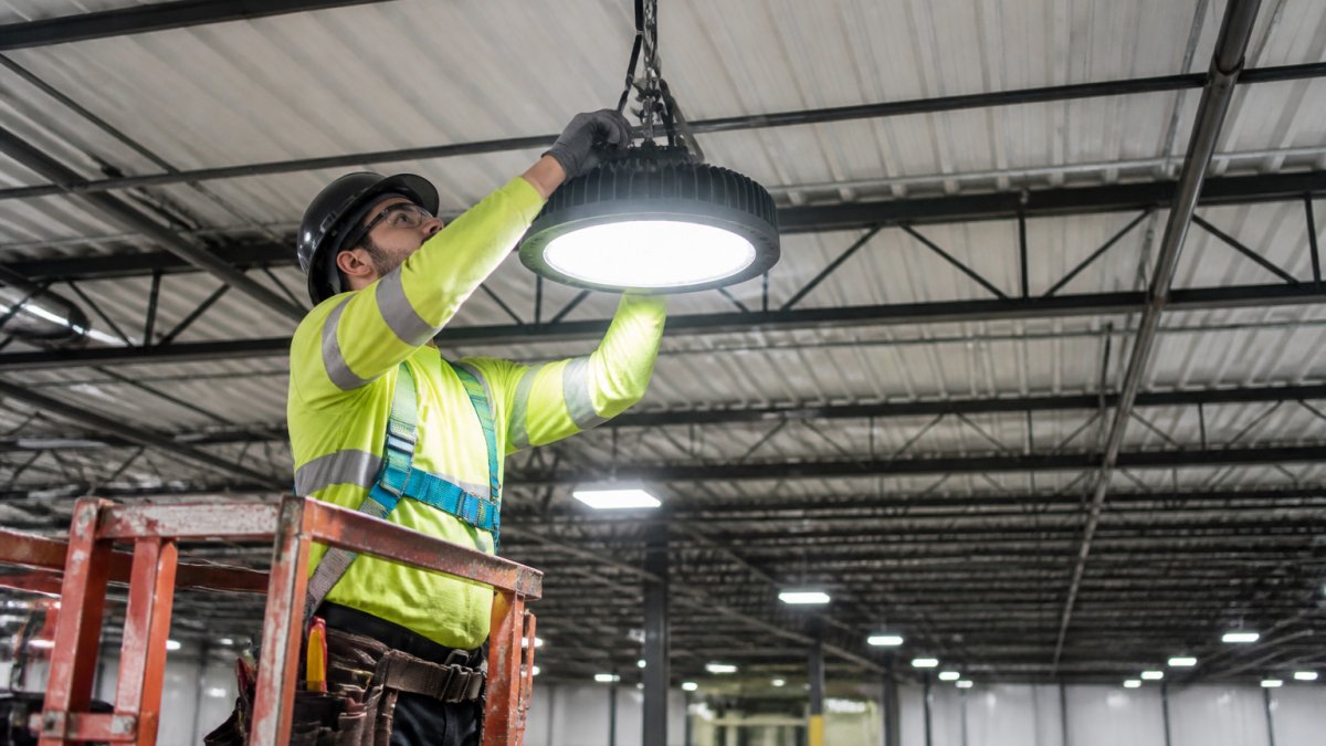 Professional electrician installing modern LED high-bay fixture in industrial warehouse ceiling, with safety equipment and proper rigging visible, showing bright white LED light output in spacious facility