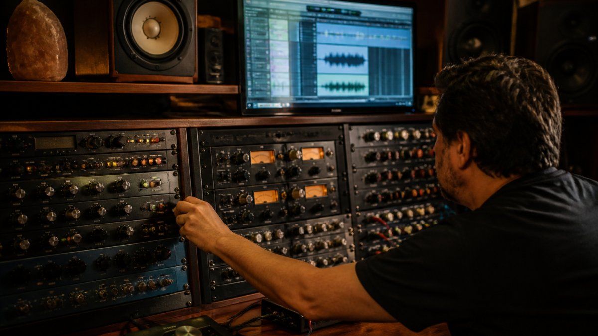 A recording engineer adjusting outboard analog gear in a studio rack while a computer screen shows a digital audio workstation with vocal tracks, warm studio lighting, cables and knobs visible