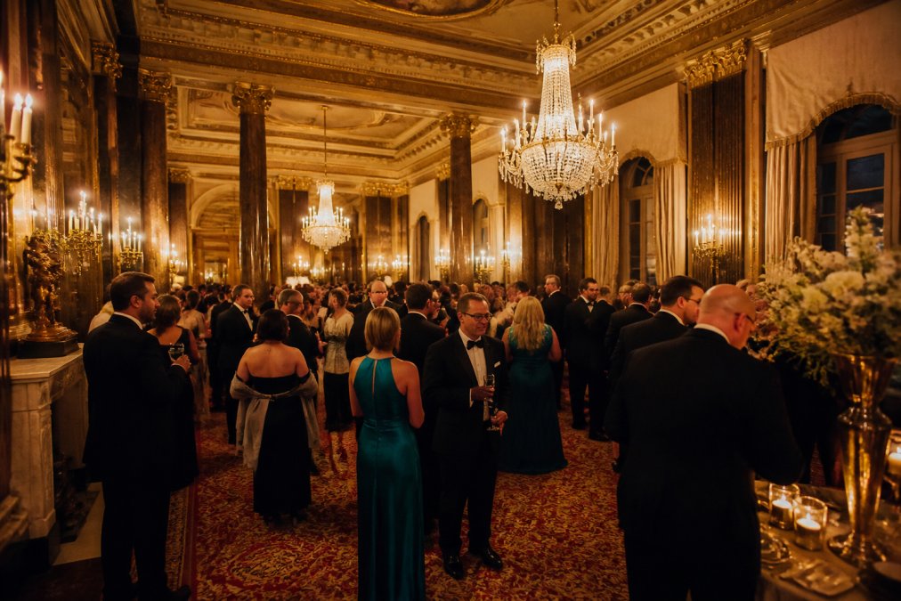 Elegant interior of Anderson House's formal reception hall with ornate gilded chandeliers, soaring ceilings, and guests mingling during an upscale event with candlelit ambiance