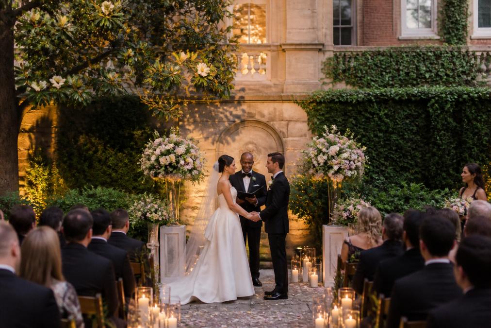 Bride and groom exchanging vows in Anderson House's historic garden setting with ivy-covered walls, pastel florals, magnolia trees, and candlelit ambiance during golden-hour light