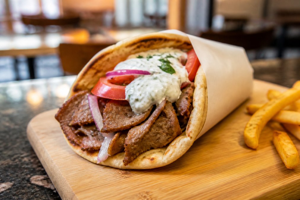 A freshly made gyro wrapped in warm pita showing layers of seasoned meat, tzatziki sauce, tomato, and onion on a wooden board in a cozy restaurant setting