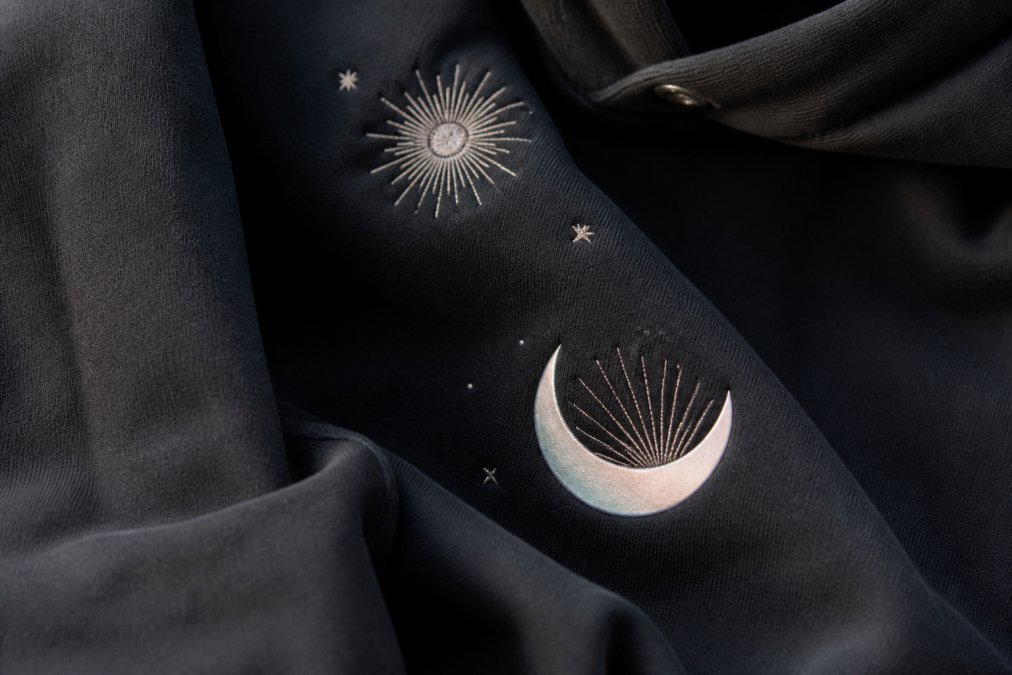 Close-up detail shot of embroidered celestial accents and reflective print on a premium oversized hoodie, showing fabric texture and design craftsmanship